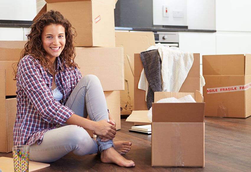 The image depicts a Hire Man and Van removal van parked on a residential driveway in Holt, with the rear doors open, revealing a large collection of waste materials inside. The waste includes several black plastic garbage bags, flattened cardboard boxes, and a few loose items such as a broken wooden chair and a discarded mattress, all appearing to be household rubbish. The van, painted in a neutral colour with visible branding on its side, is positioned on a paved driveway that extends towards a suburban street, with a well-maintained garden area on the left. The surrounding environment shows a typical residential setting, with a fence and some shrubbery partially visible behind the van. The scene is illuminated by natural daylight, diffused through overcast skies, creating soft shadows around the vehicle and waste. The overall composition illustrates a professional rubbish removal process, with Hire Man and Van prepared to load and dispose of the waste efficiently, supporting local waste management services in the Holt area with a focus on domestic clearance.